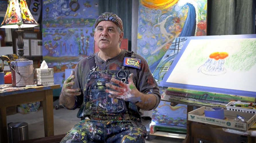 An instructor is sitting down in an art studio talking with his hands to the camera during an interview.
