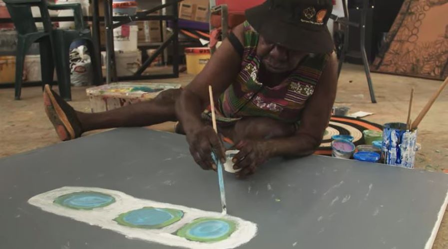 An older adult woman from an Aboriginal community controlled art centre lays down on the floor painting a large canvas.