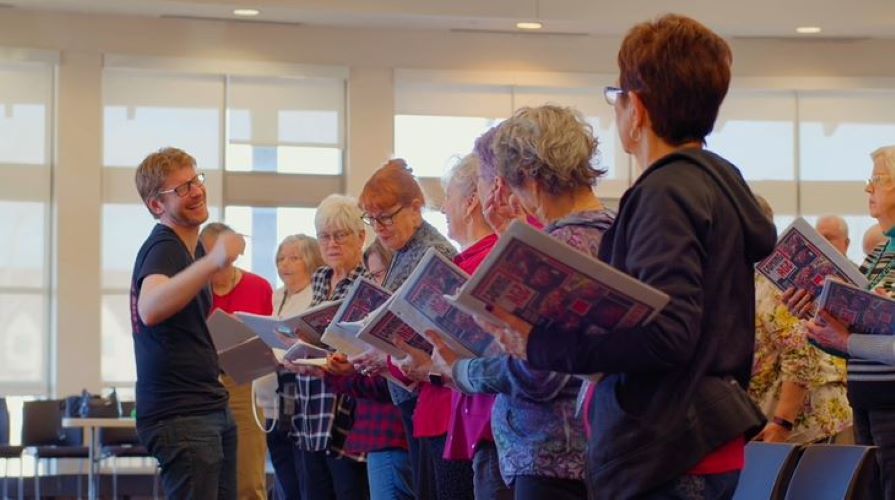 A group of older adults sing together while an instructor conducts them. The instructor is smiling and laughing.
